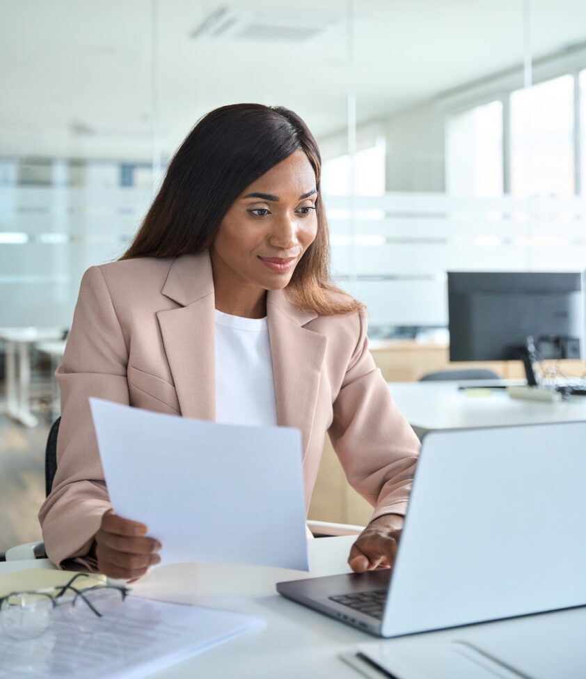 Female account manager, professional busy African American business woman executive holding document using laptop checking online corporate financial data working sitting at office desk, vertical.
